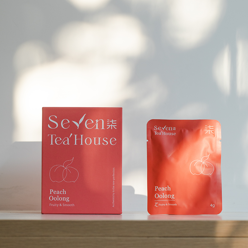 A redish pink box and packet of peach oolong tea bags sitting on a wooden surface with a white background